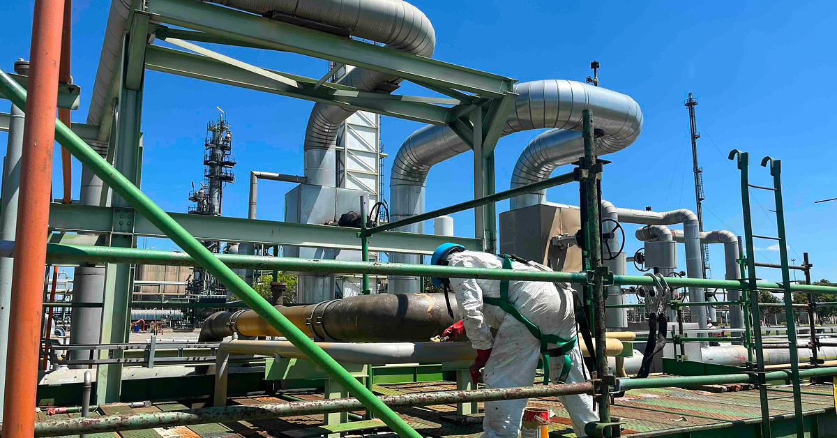 Worker in safety gear securing scaffolding at an industrial site, preparing for painting among large metal pipes and structures.