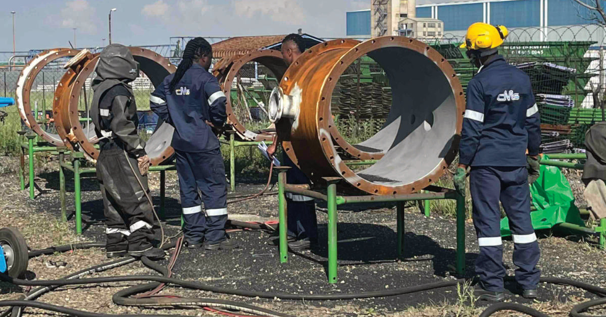 Workers in protective gear are inspecting large metal pipes outside, with industrial equipment visible in the background.