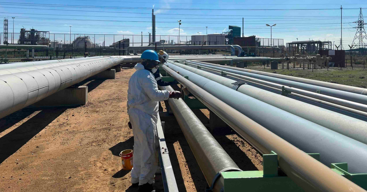 A person in protective gear working on industrial pipes outdoors on a sunny day.