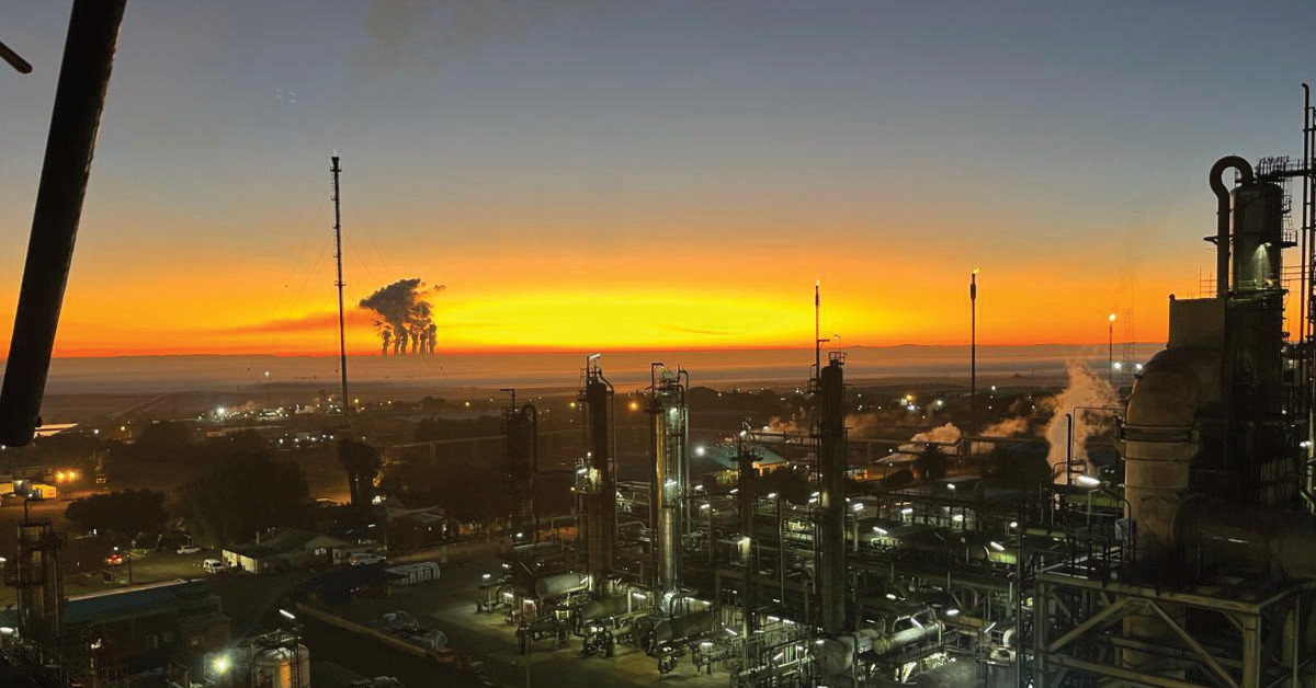 An industrial refinery against a bright orange sunset, with dark smokestacks releasing steam.