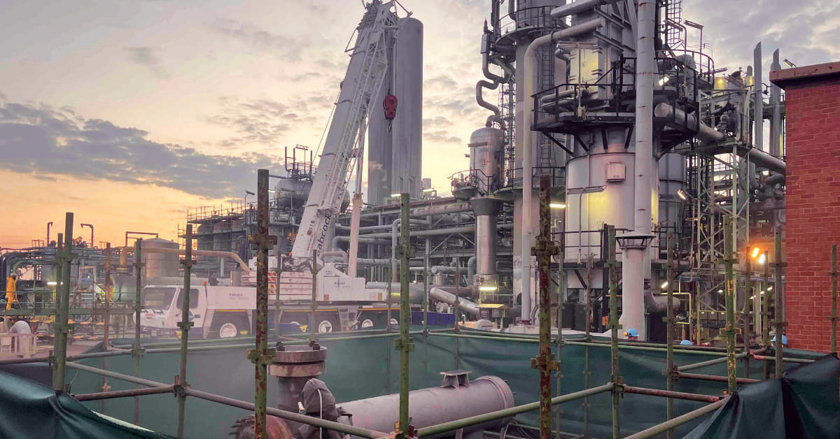 An industrial plant with metal structures and cranes, set against a sunset backdrop, surrounded by scaffolding and green barriers.